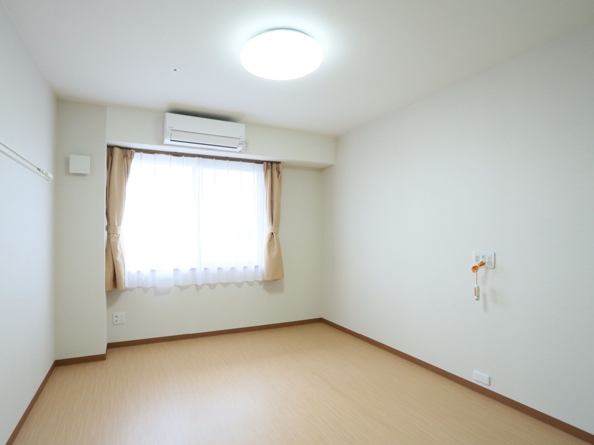 日当たりの良い居室で、窓にはカーテンとレースカーテンが付けられている。壁にはコンセントがあり、ナースコールが設置されている。