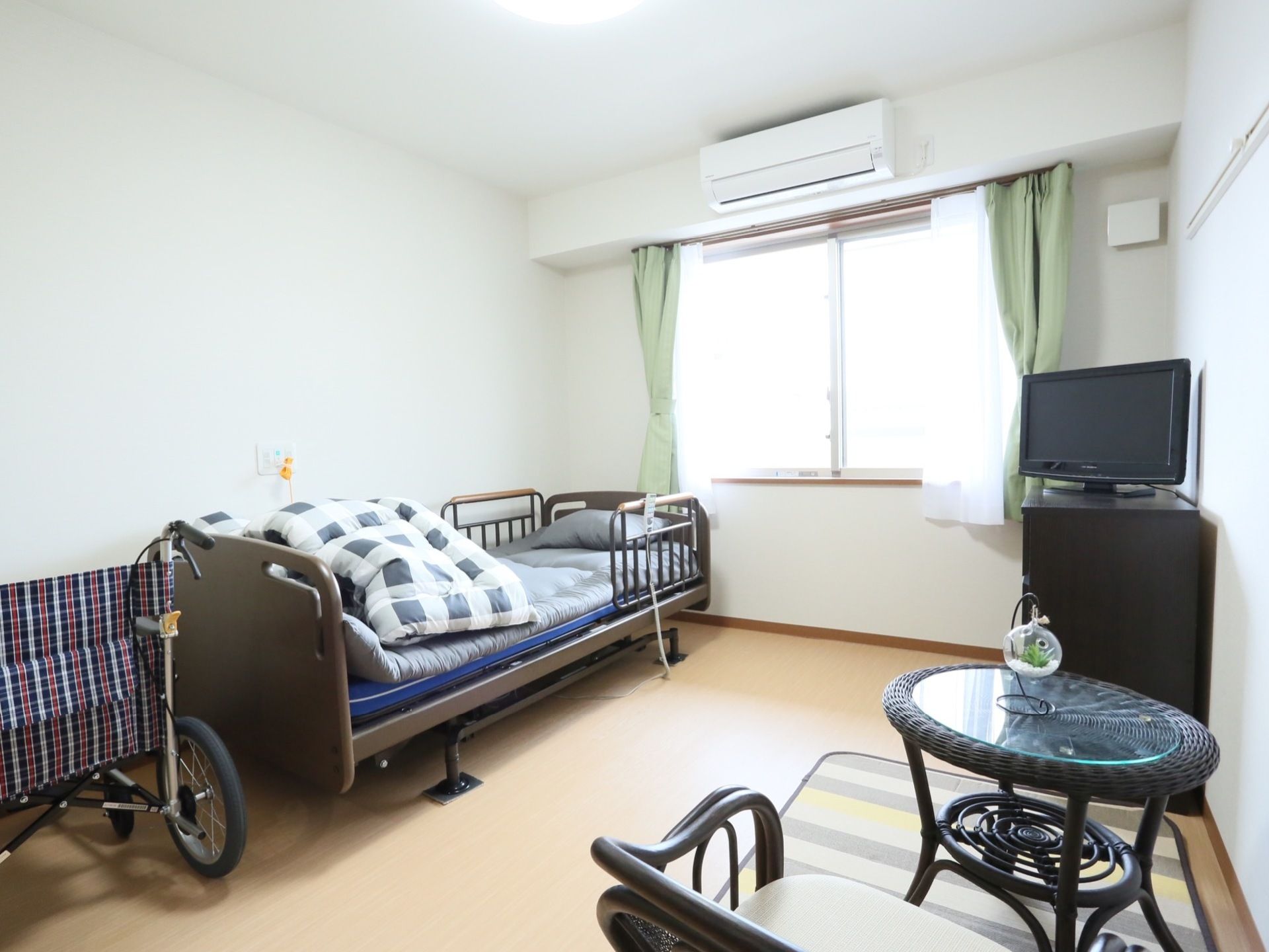 日当たりの良い窓のある居室。壁際には介護用ベッドがあり、棚の上にはテレビが置かれている。ラグマットの上には籐製のテーブルセットがある。