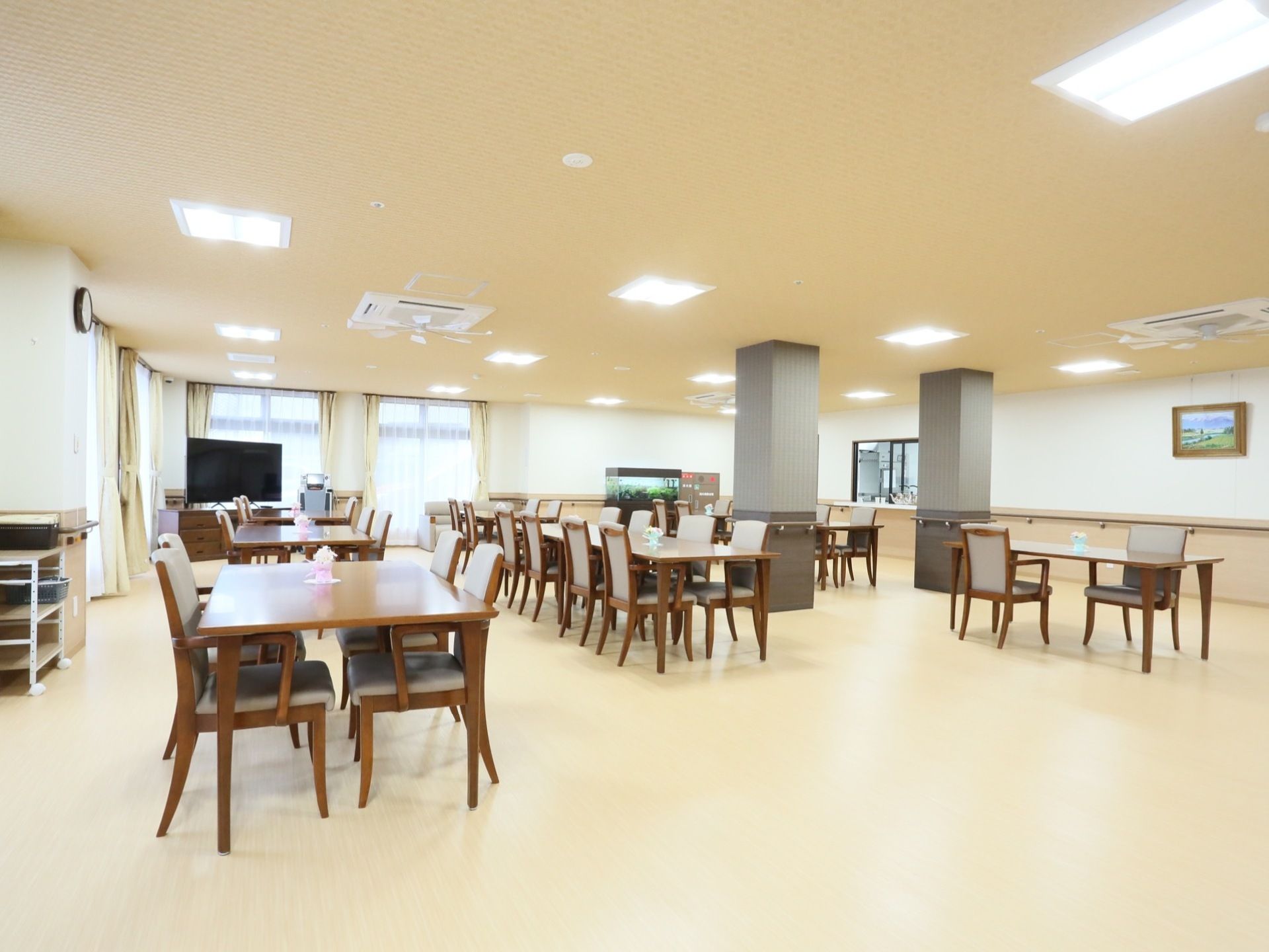 床と天井がクリーム色で、広く明るい食堂。テーブルと椅子がたくさんあり、壁際には大きな窓が２つ並んでいる。壁には額縁に入った絵が飾られている。
