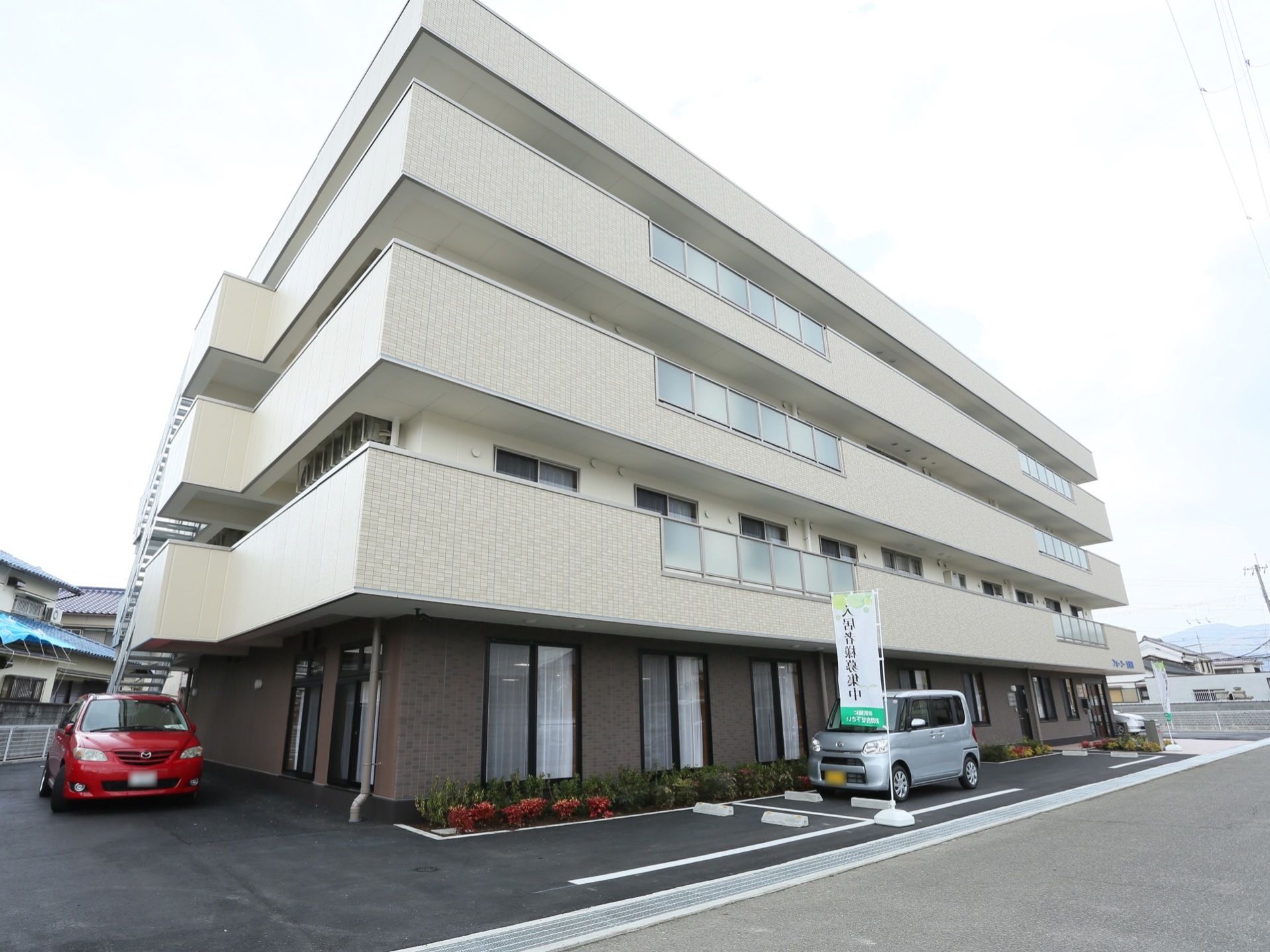 下部分が茶色、上部分が白いタイル張りの４階建ての大きな建物がある。外付けの非常階段が設置されている。手前と横部分は駐車スペースになっている。