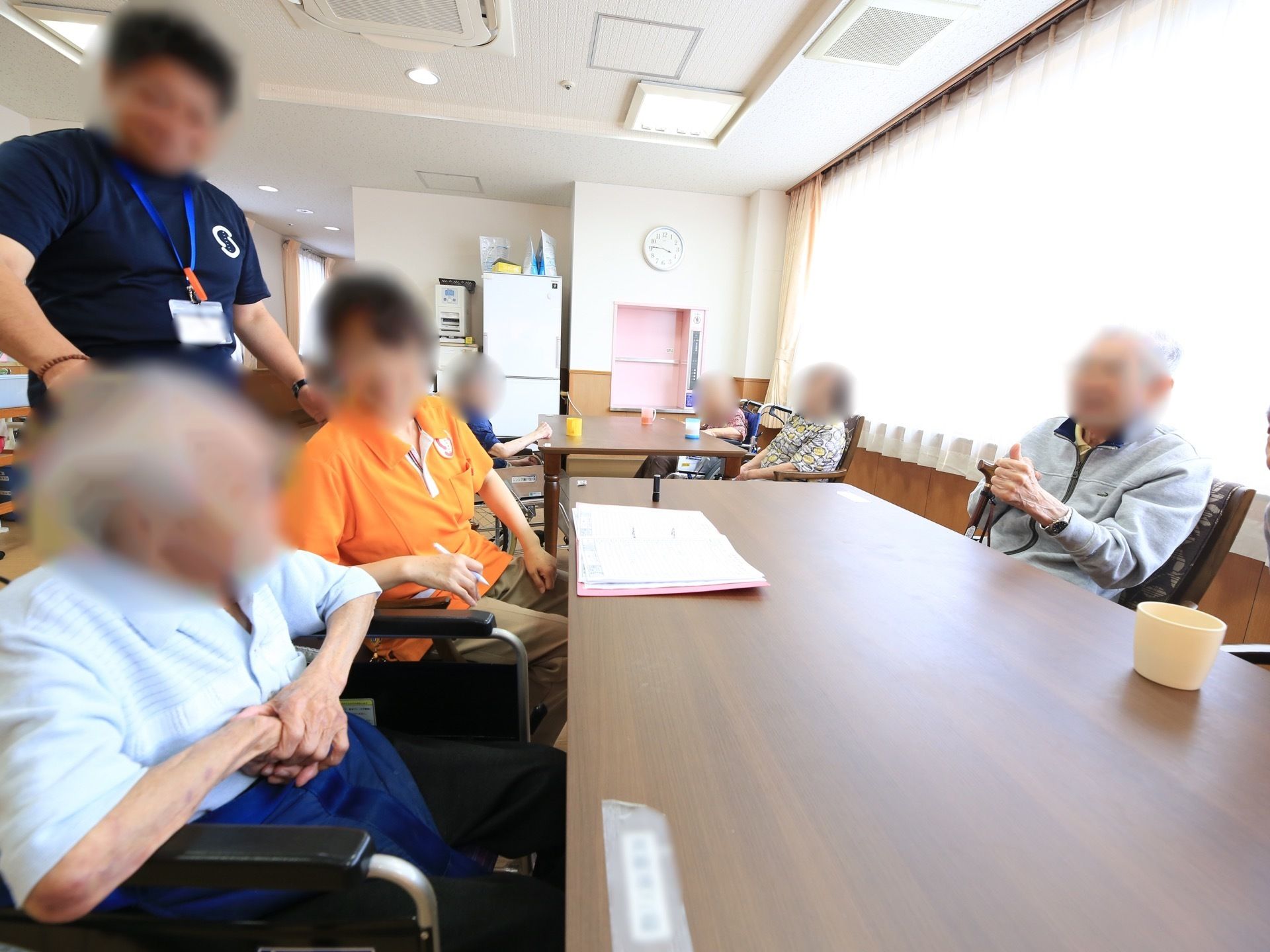 共同生活室のテーブル席で、入居者様がスタッフと談笑している。みんなが笑顔で、和やかで楽しそうな雰囲気。