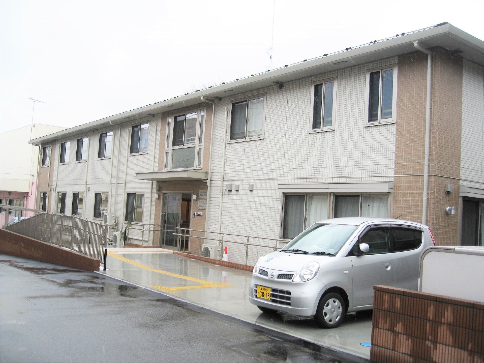 横に長い２階建ての建物はベージュとブラウンで、玄関先には屋根が付き、駐車場もある。