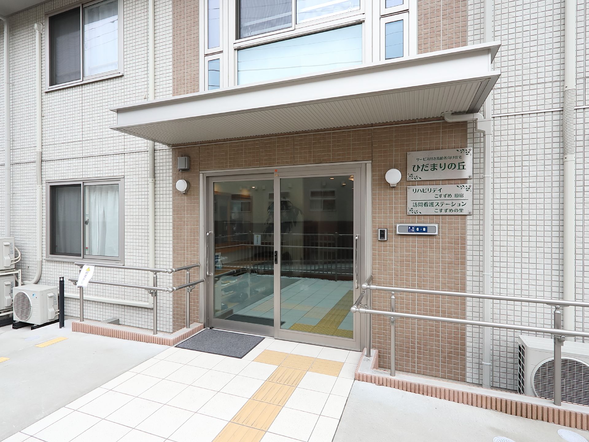 玄関横の外壁には表札やインターホン、照明が設置されている。地面にはグレーのマットが敷かれており、黄色の点字ブロックが埋め込まれている。