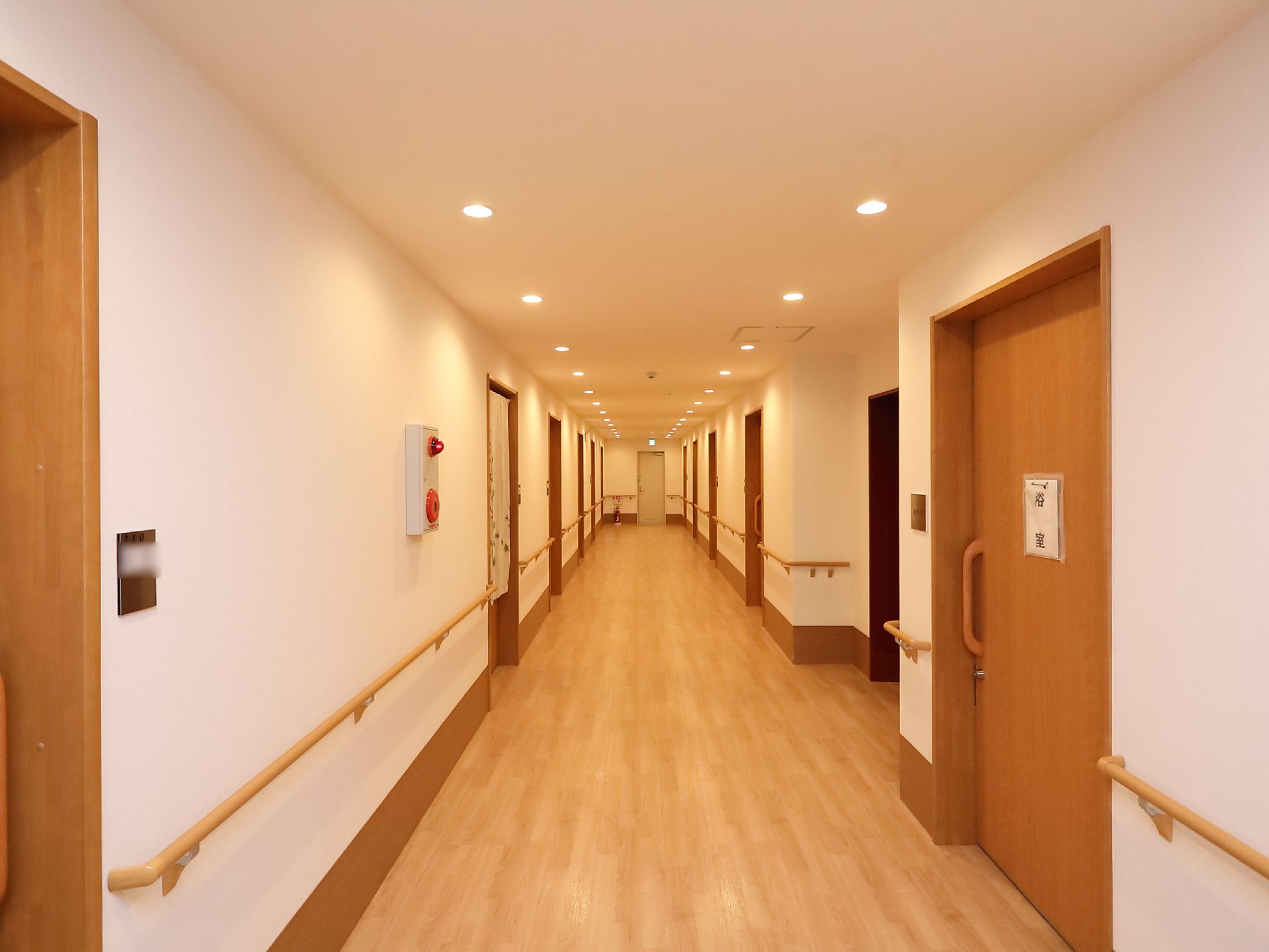 横幅のある広々とした廊下で、壁には手すりが取りつけられている。途中に火災報知器が設置されており、奥には消火器が置かれている。