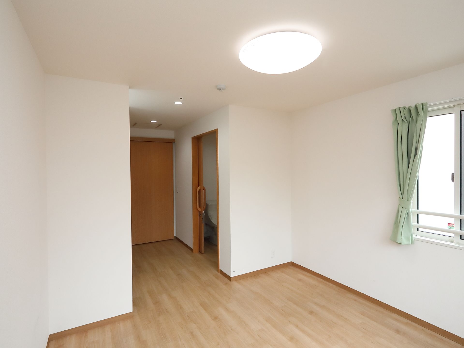 居室の通路部分にトイレが設置されている。窓には手すりが設けられており、緑色のカーテンが取りつけられている。