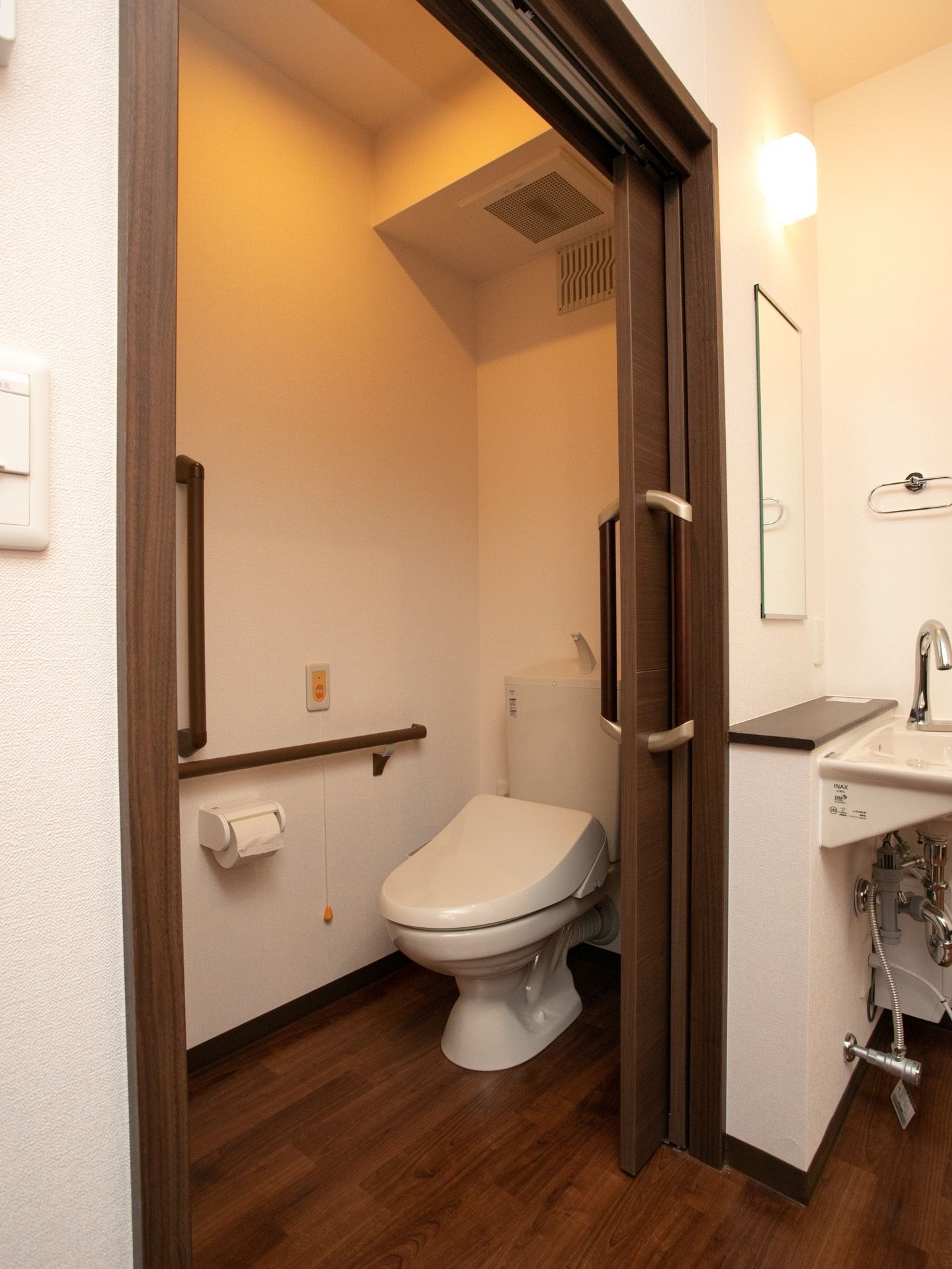 木製の引き戸の奥にトイレがある。壁に緊急通報装置が付いている。トイレを出たところには洗面台が設置されている。