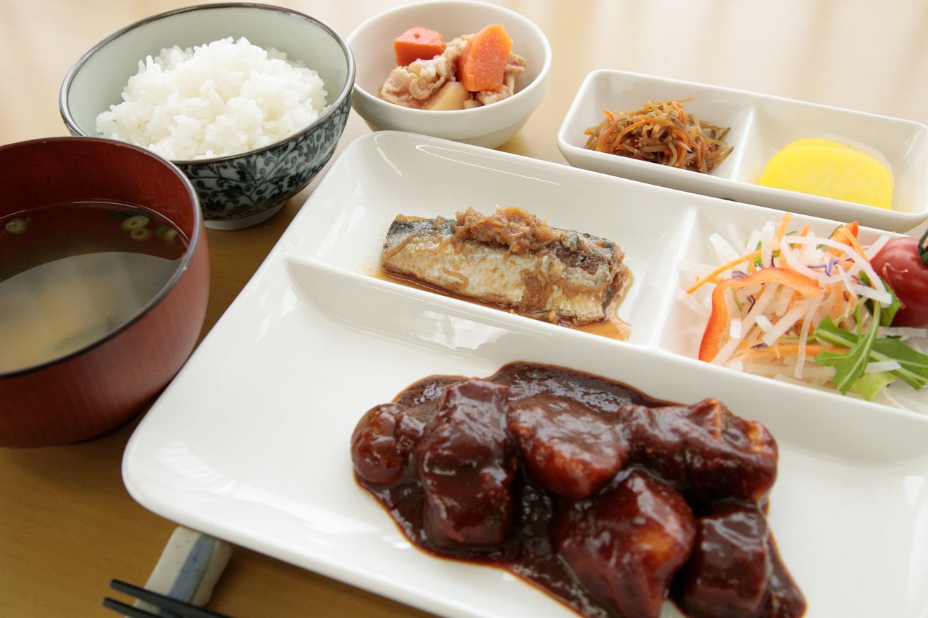 テーブルの上に黒いお盆が置かれ、その上にご飯、みそ汁、酢の物、炒め物、煮物などバランスの良い献立が並んでいる。