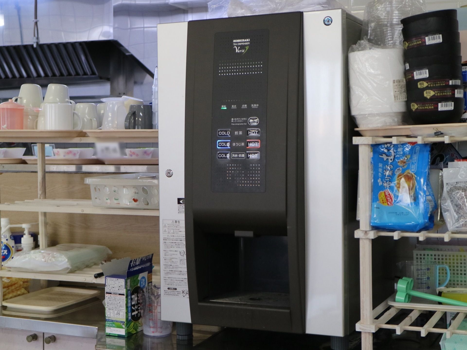 食堂に設置してある煎茶、ほうじ茶、温冷水の給茶機である。そばの棚には、お茶、青汁などや入居者様のコップ類が置いてある。