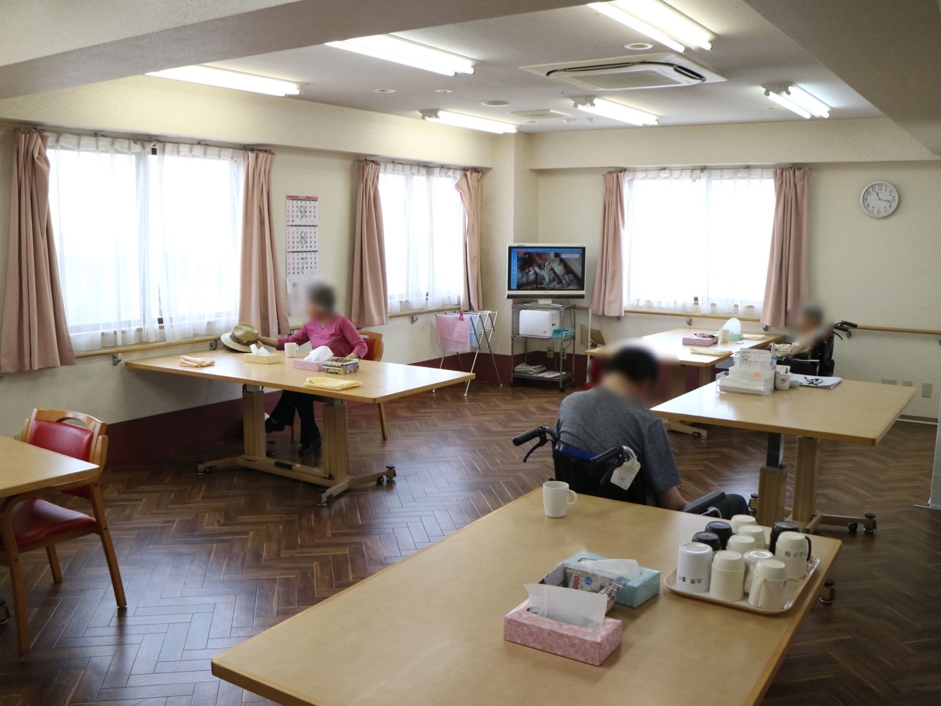 食堂にはテーブルが5卓あり、入居者様が椅子や車椅子で座っている。奥にはテレビがあり、周りの窓から明るい日差しが入っている。