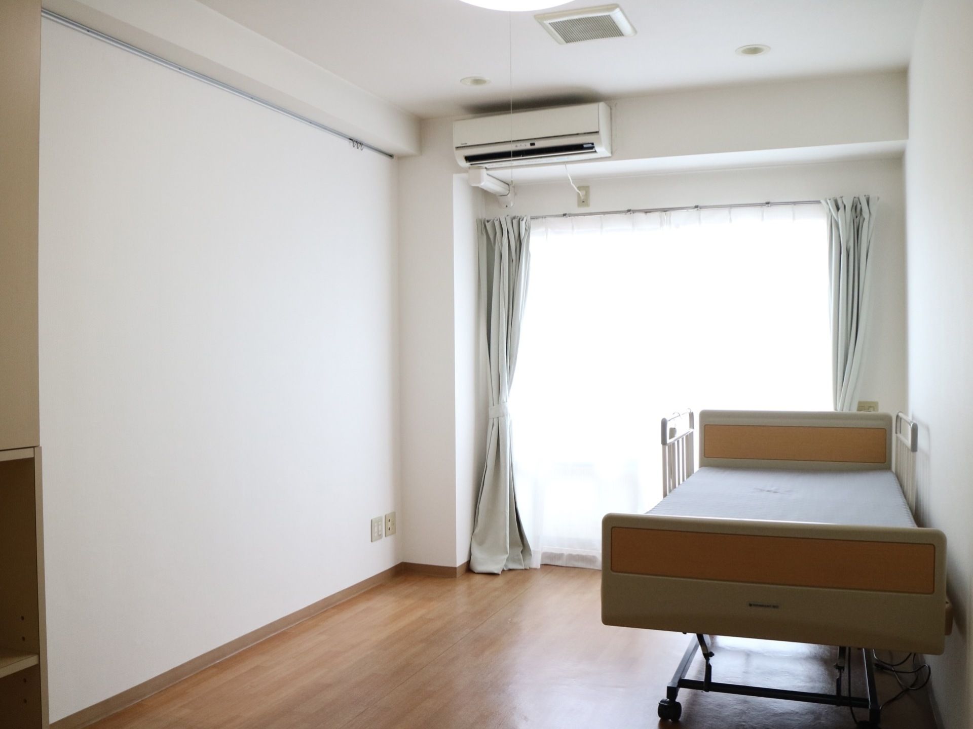 居室内部はフローリング仕様である。掃き出し窓には淡い色のカーテンが掛かっており、ベッドとエアコンが設置されている。