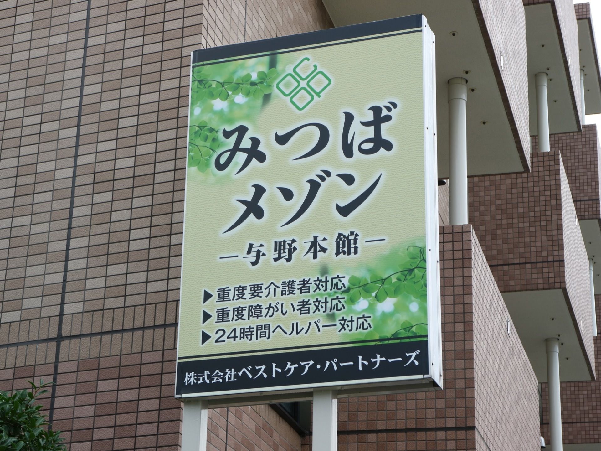 建物の外に設置されている施設の看板である。緑色を基調とし、大きく施設名と対応できる入居者を記してある。運営会社の名称も入れてある。