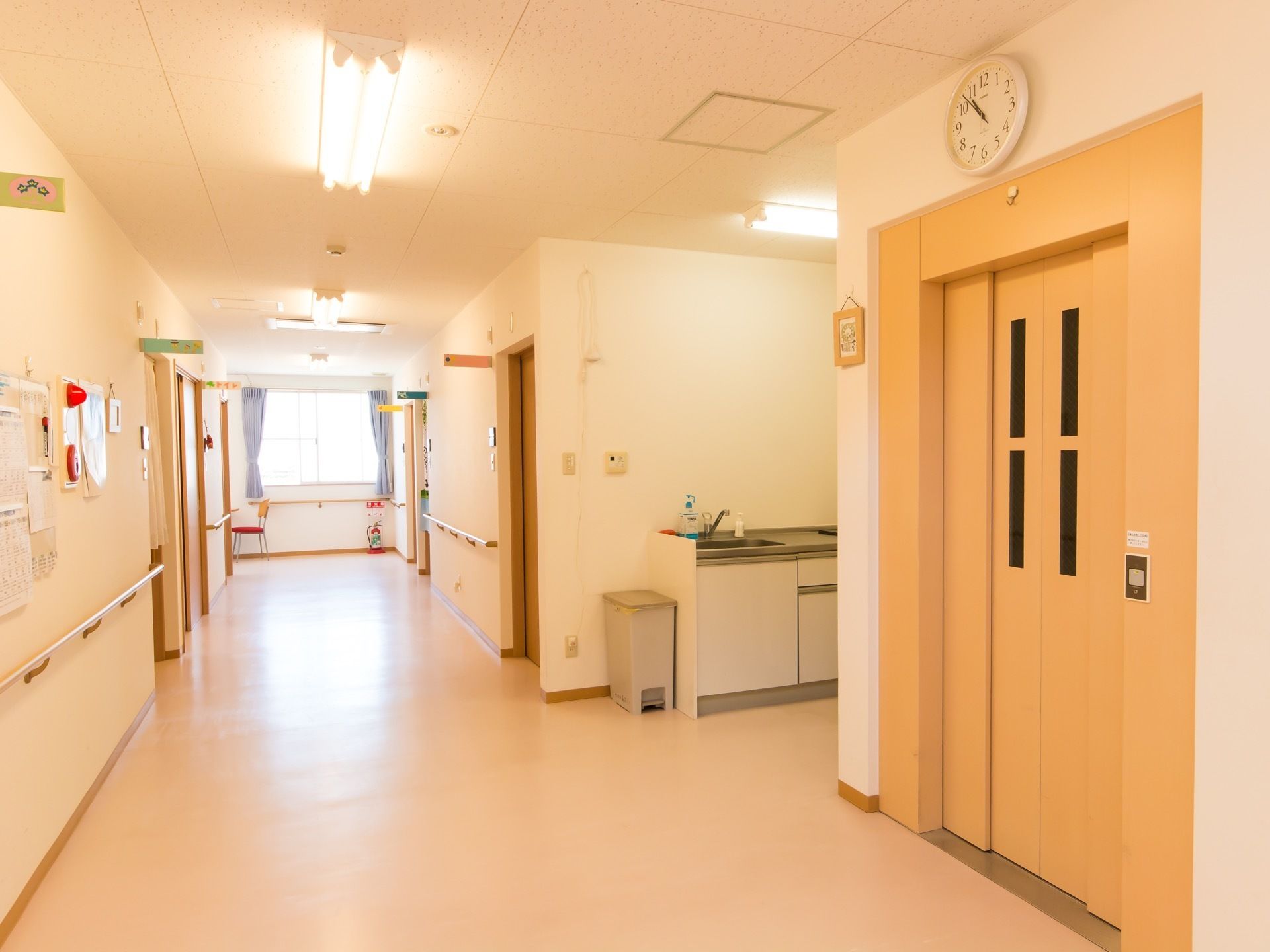 クリーム色の明るい床の廊下が続いている。エレベーターが設置されていて、その奥には流し台が見える。壁には手すりがついている。