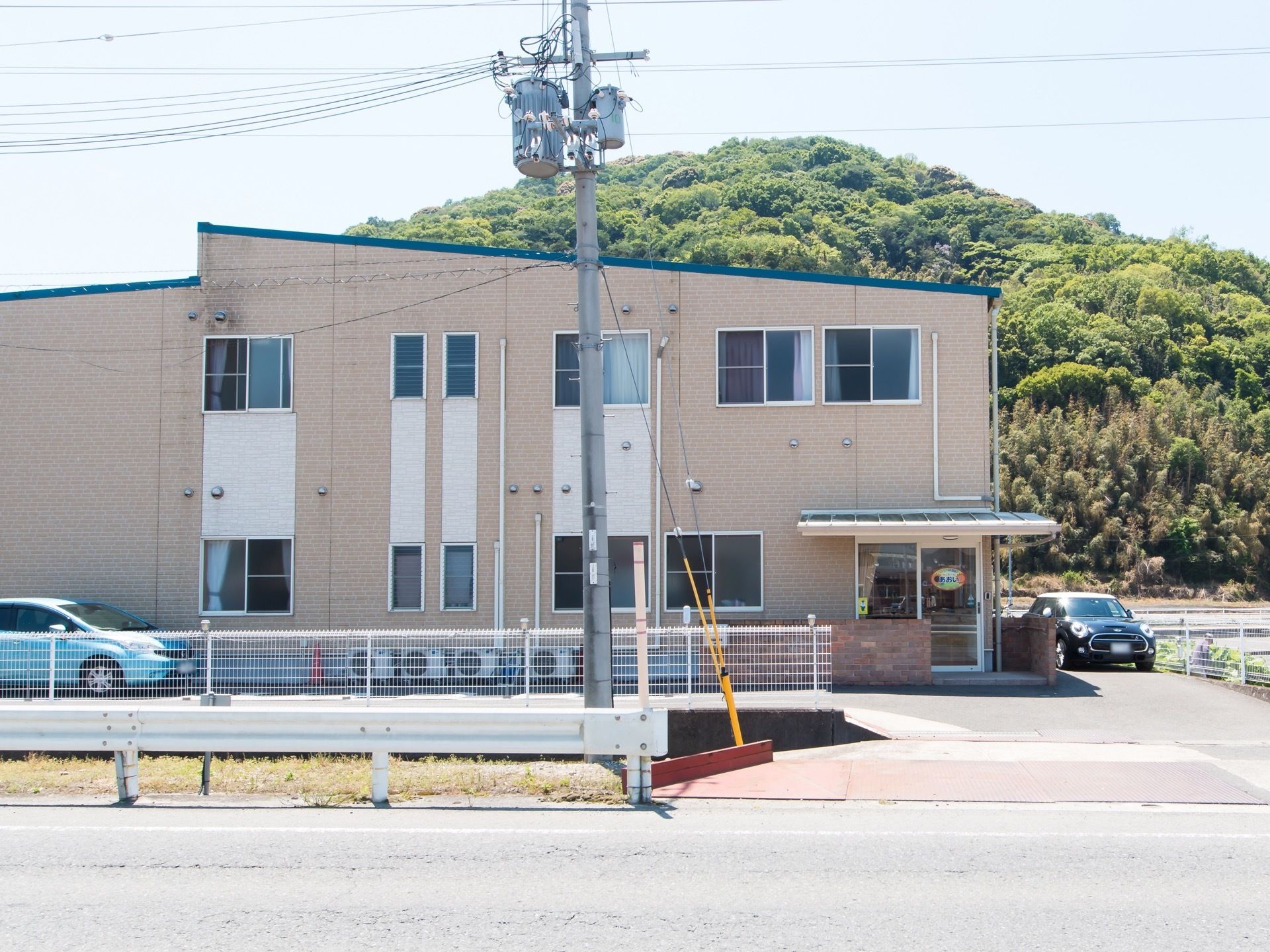 車道から歩道を挟んだ敷地に2階建ての建物がある。入口は自動ドアになっていて、建物の脇に車が数台停まっている。