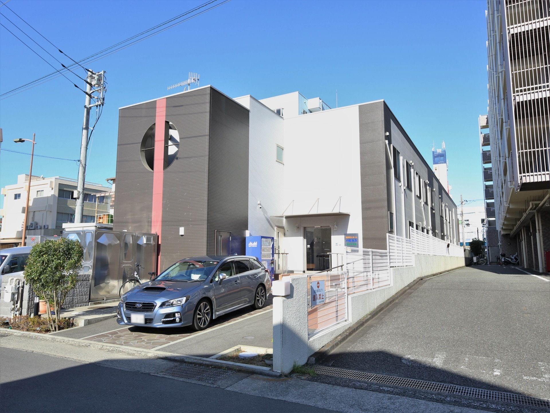 敷地内にはモダンな施設の建物があり、玄関前が駐車スペースになっている。玄関脇には飲料の自動販売機が設置されている。