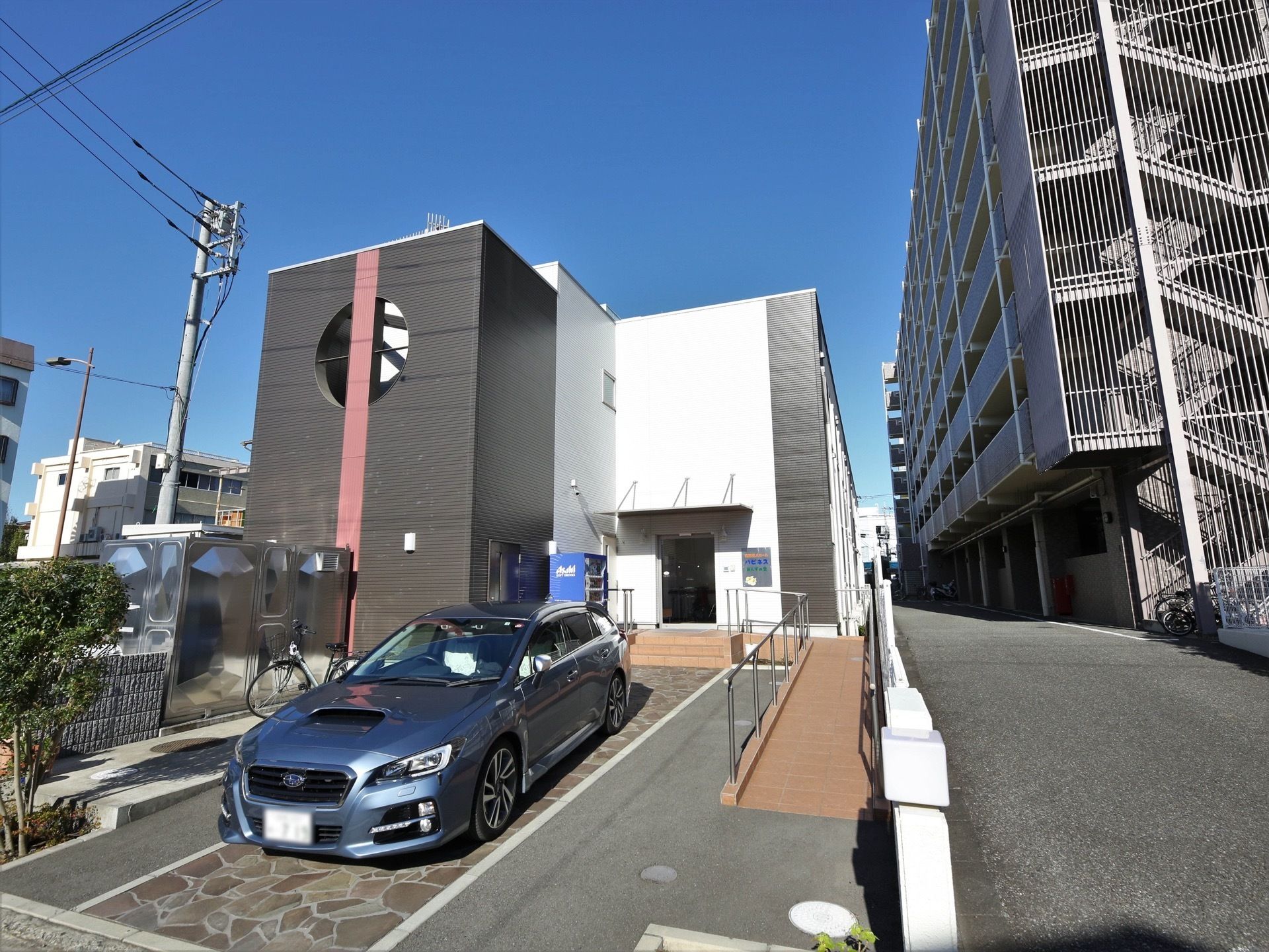 隣に大きいマンションが建っているが、施設の日当たりは良い。玄関前の駐車スペースに車が１台停まっている。