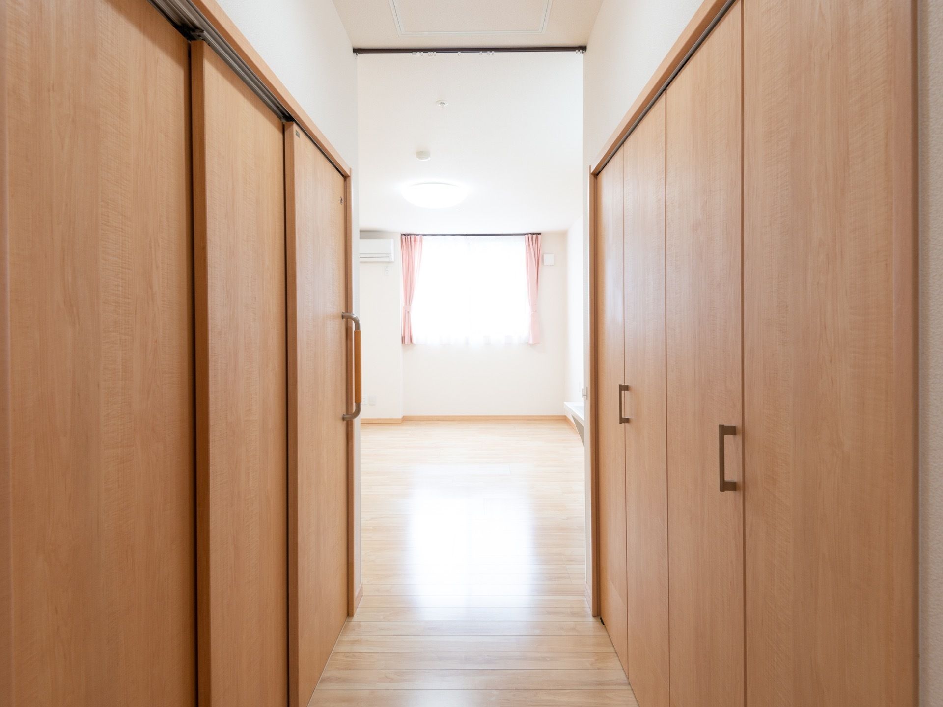 居室の入り口より、部屋の中をのぞいた様子。入り口の両側に収納庫やトイレの木製ドアがある。正面の窓から陽光が差し込んでいる。