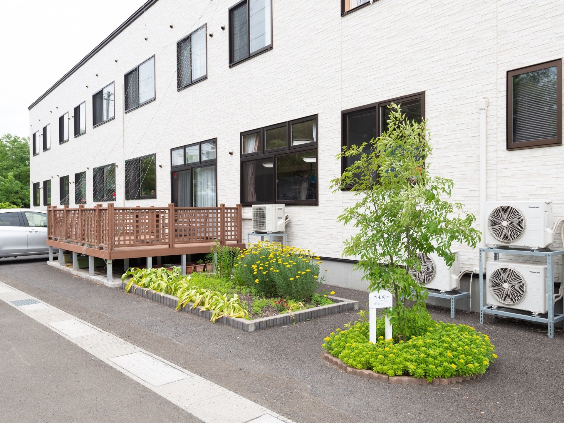 施設は白い壁の２階建ての建物となっている。ウッドデッキが設けられている。建物脇に、草花や庭木の植栽された、花壇がある。