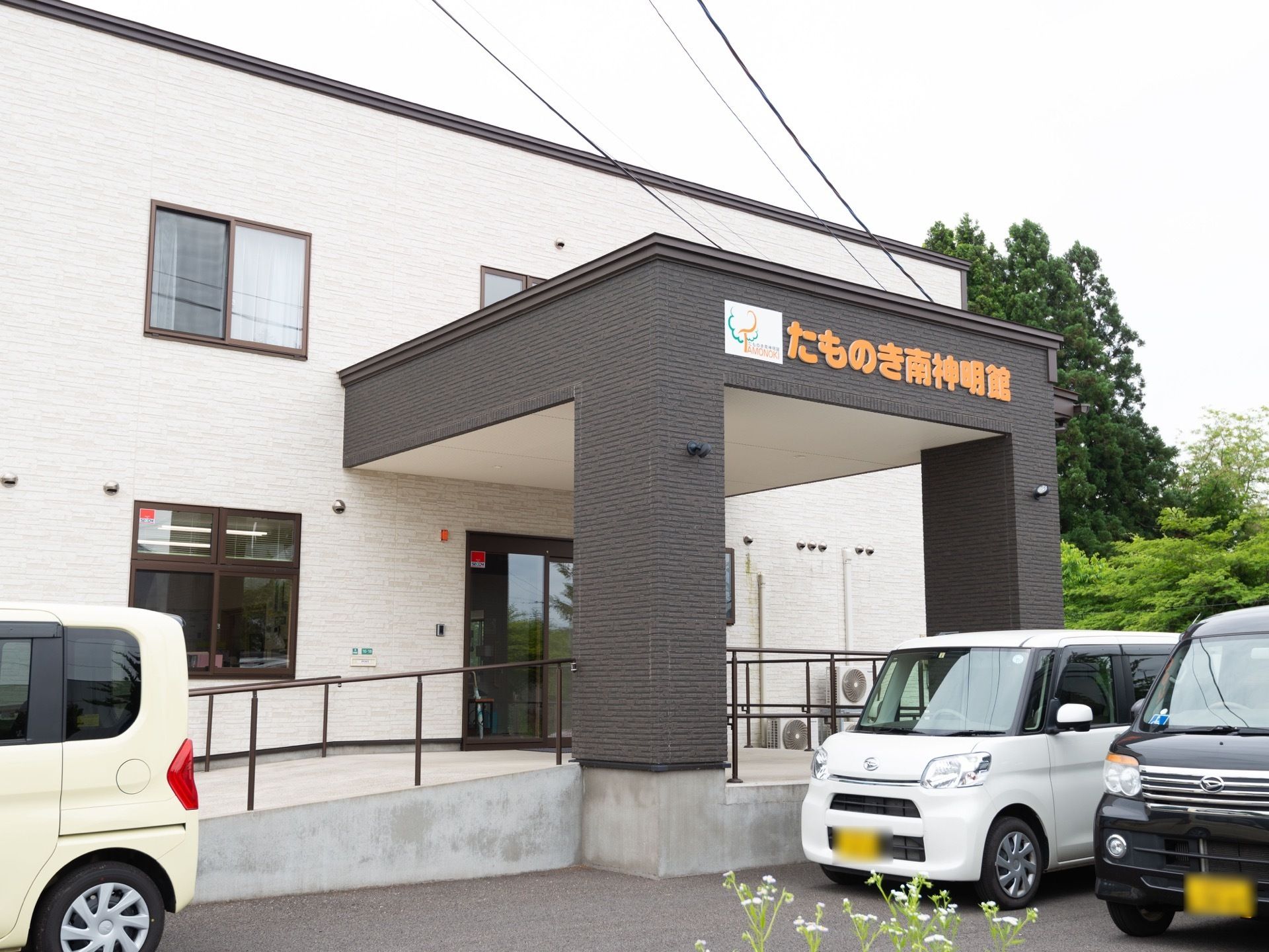 施設は白い外壁の２階建ての建物である。玄関から、ひさしが突き出している。建物前が駐車場となっており、数台の車が停められている。