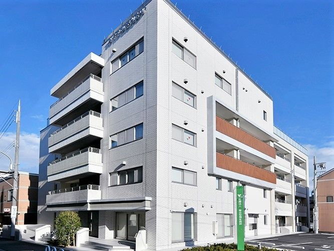 白い５階建ての建物。各フロアにベランダ付き、駐車場には緑の看板も設置されている。