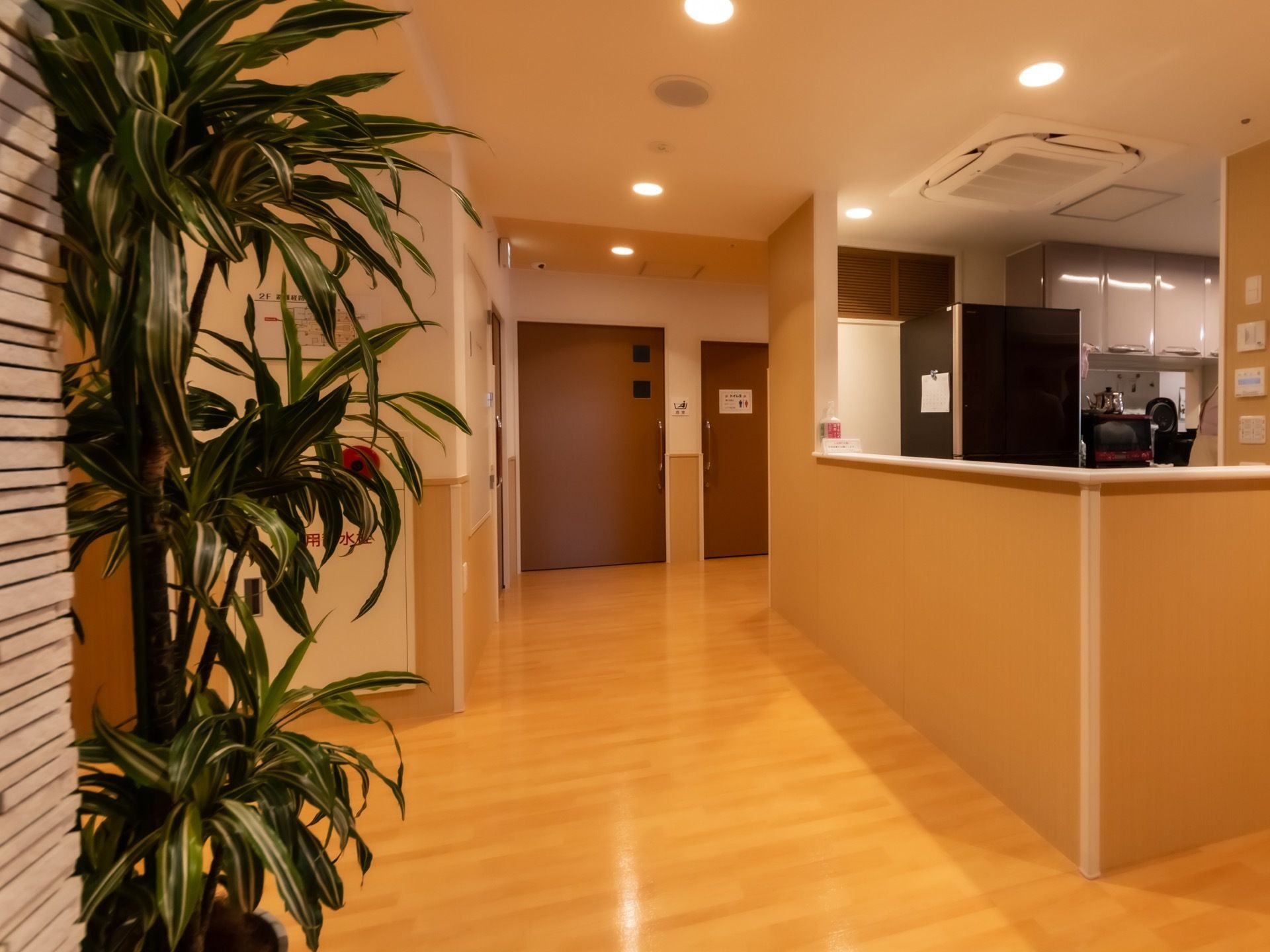 木のぬくもりが感じられる廊下で、天井には等間隔で照明が取り付けられている。キッチンの中には、冷蔵庫や電子レンジが設置されている。