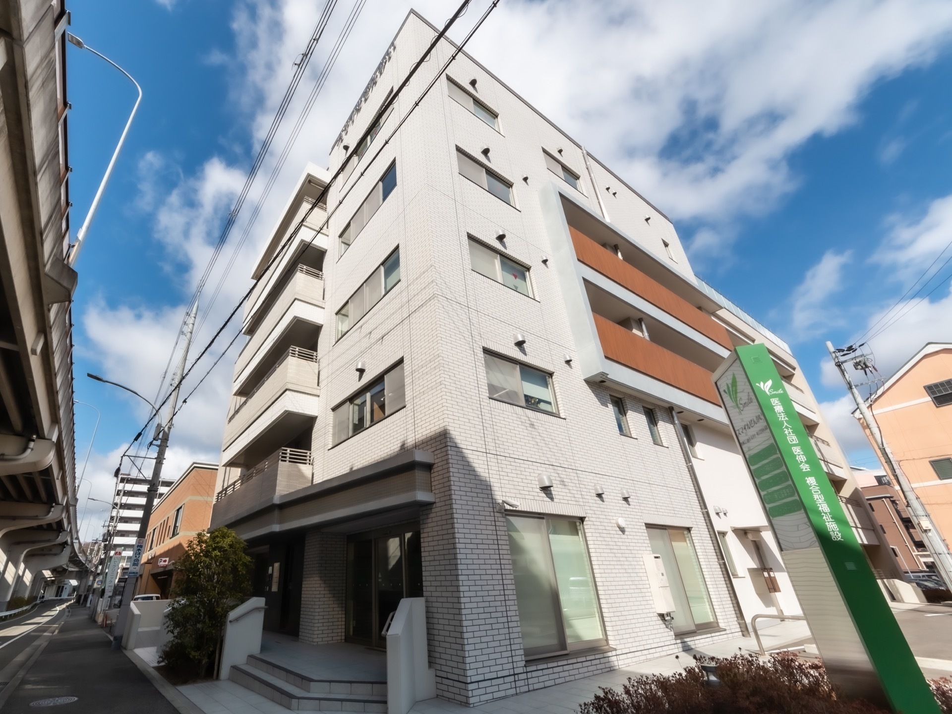 施設周辺にたくさんのマンションが建てられている。建物前には花壇が設けられており、看板が設置されている。