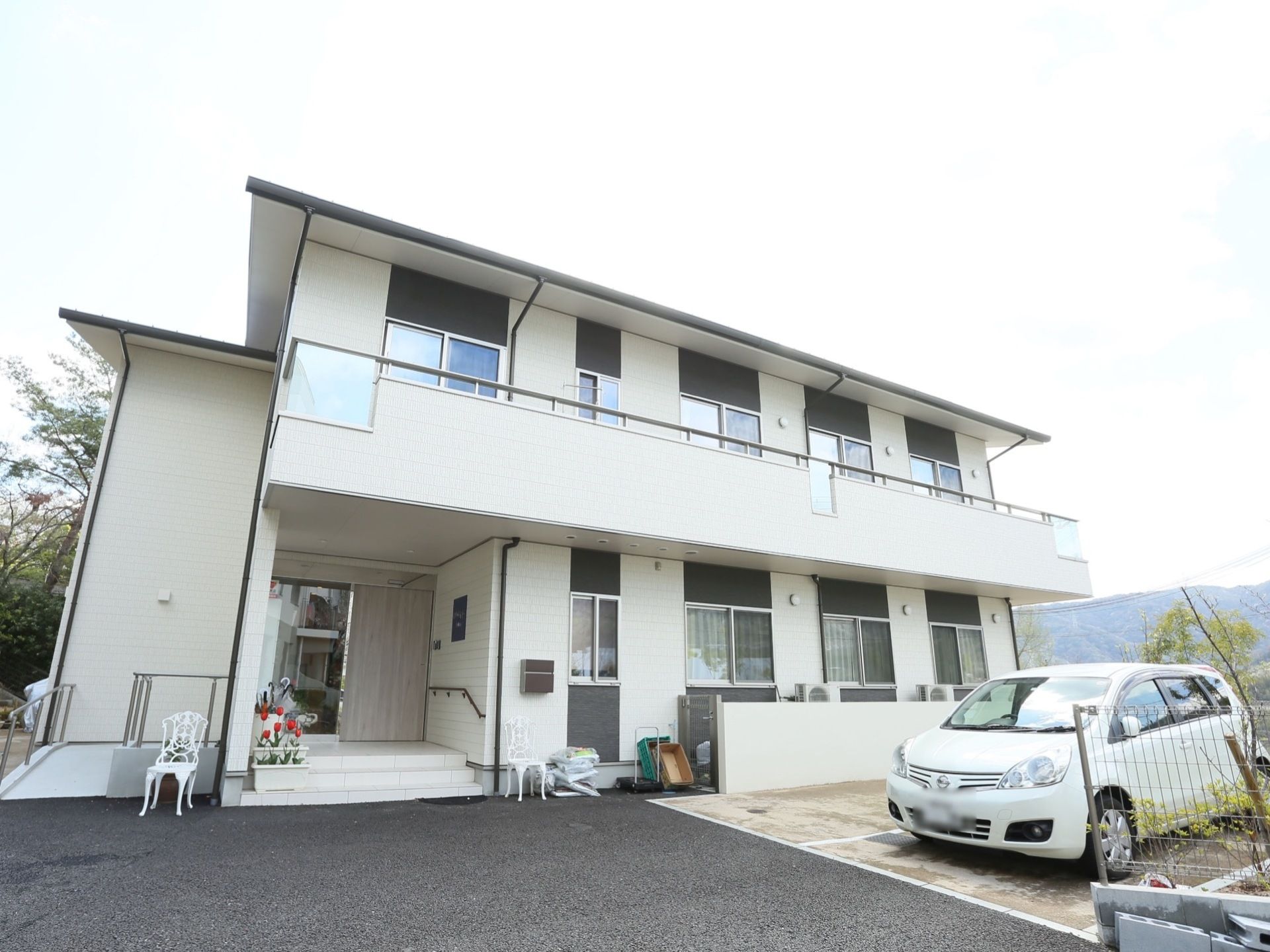 白を基調とした施設外観である。２階にはベランダが設けられている。敷地内には駐車スペースが用意されており、車が１台停まっている。