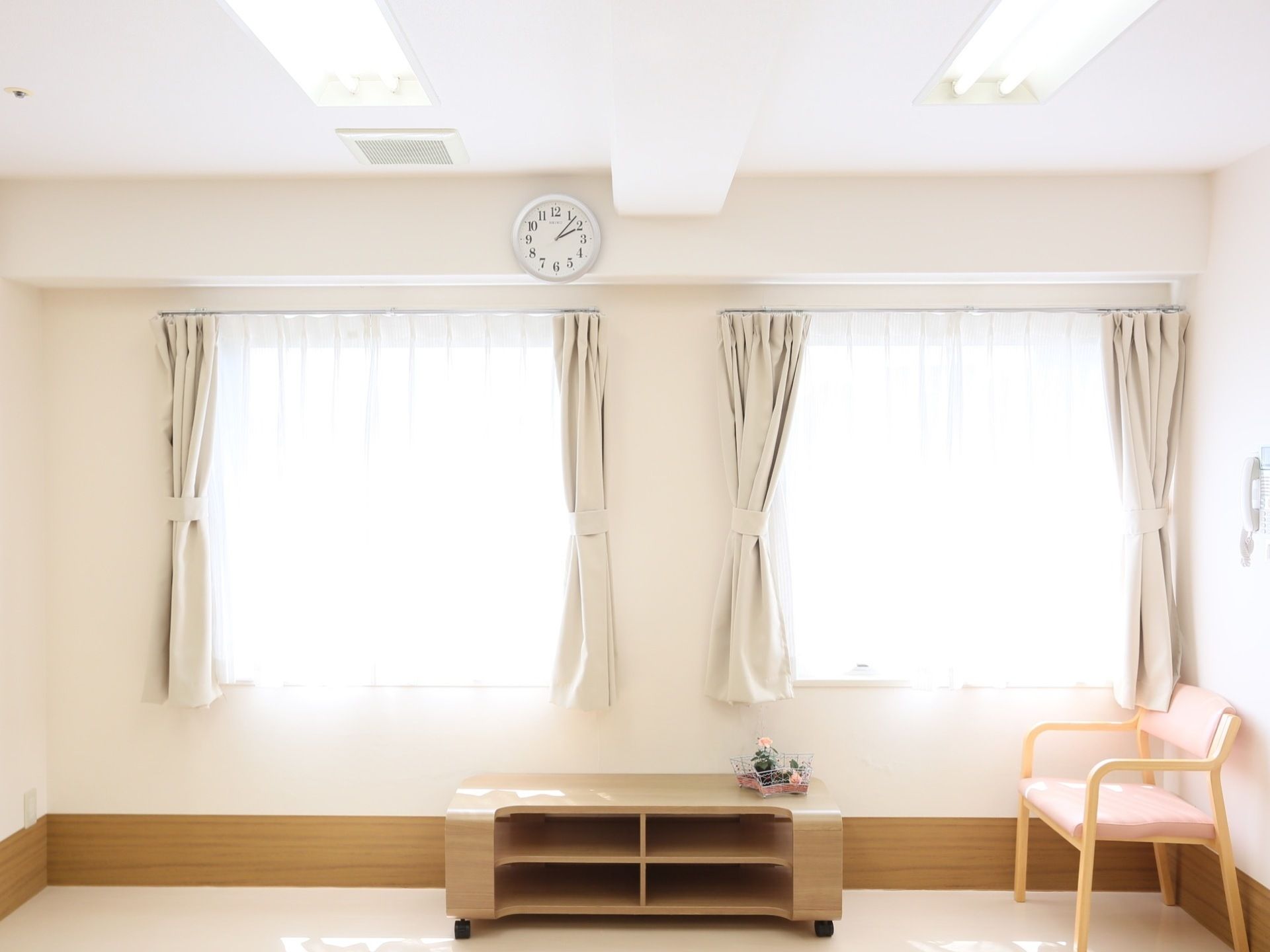 カーテンがかけられた窓が２つあり、明るい日差しが差し込んでいる。ピンク色の椅子とキャスター付きの台が置かれている。