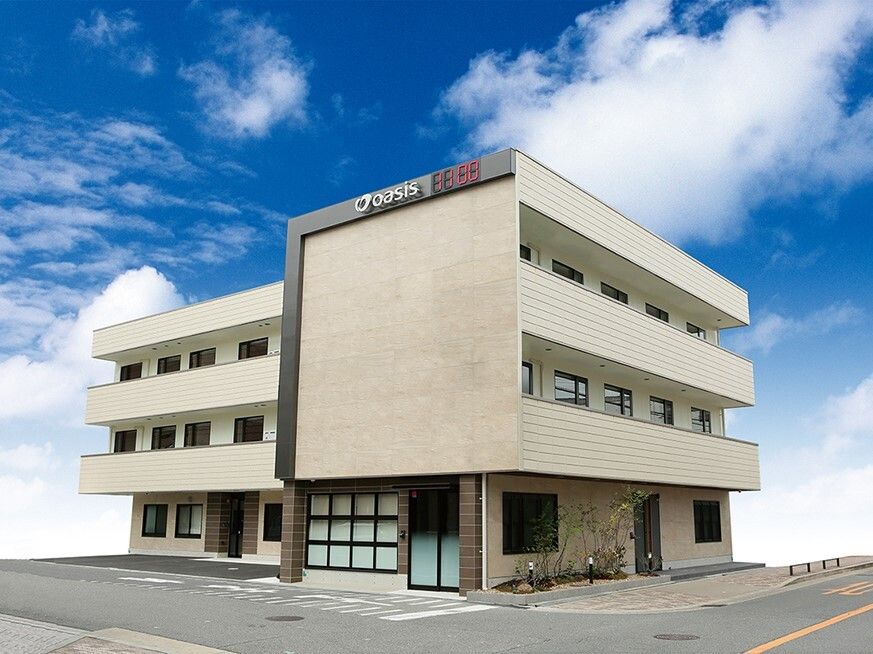 3階建ての建物はベージュ系で外壁には施設名が書かれている。各フロアにベランダがあり、玄関横には草木が植えられている。