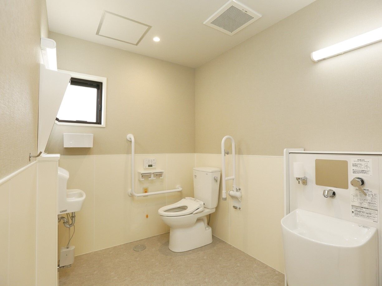 広いトイレは白を基調としていて、トイレの両サイドには手すりが付き、オレンジの呼び出しボタンも設置されている。