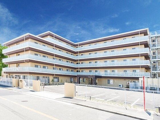 ベージュの外壁の５階建ての建物はエル字型で窓がたくさん付いている。ベランダや屋外階段も付いている。