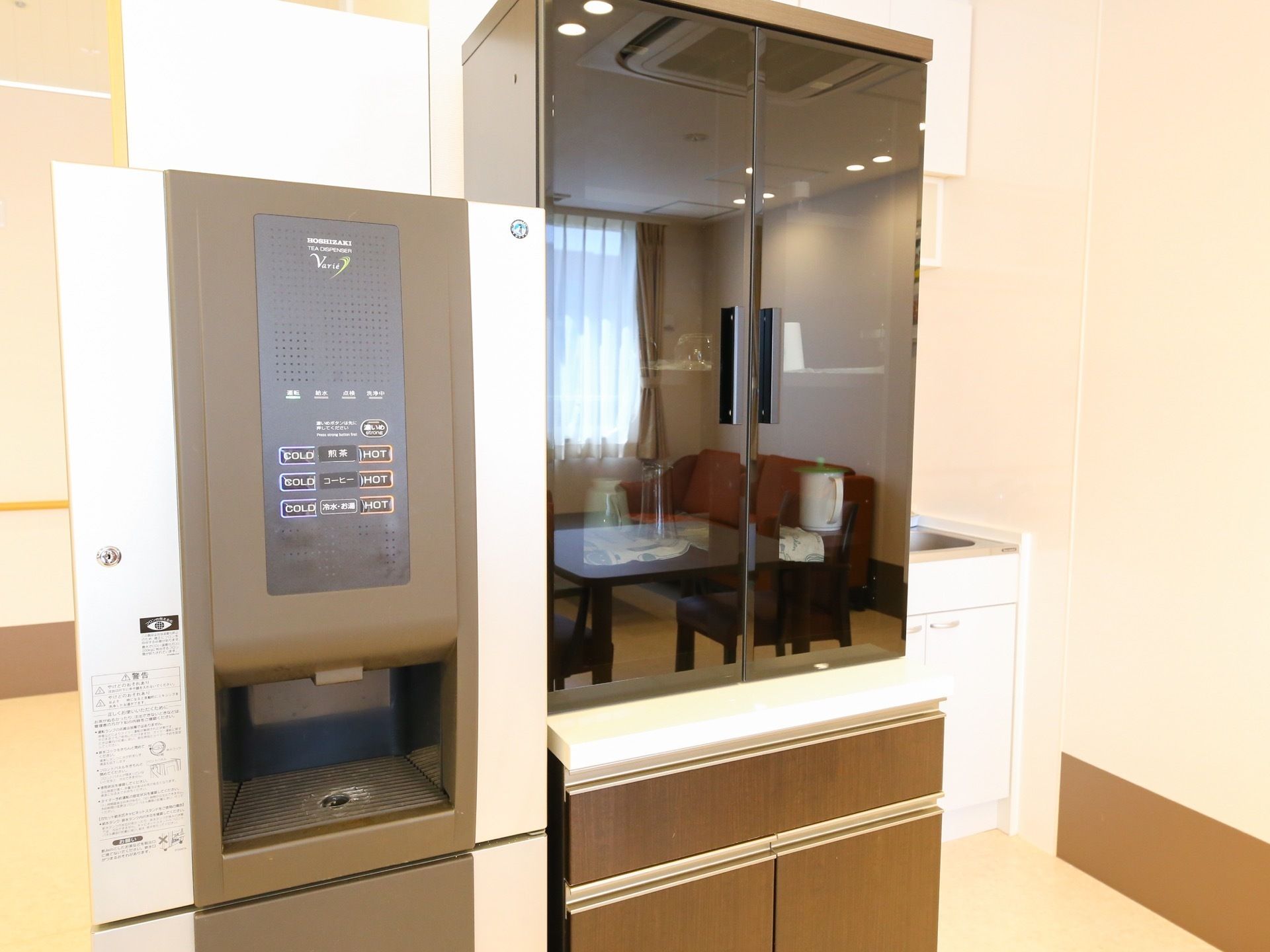 黒い鏡面仕上げの扉の食器棚があり、その横に給茶機が置かれている。棚の奥には、キッチンシンクが見える。