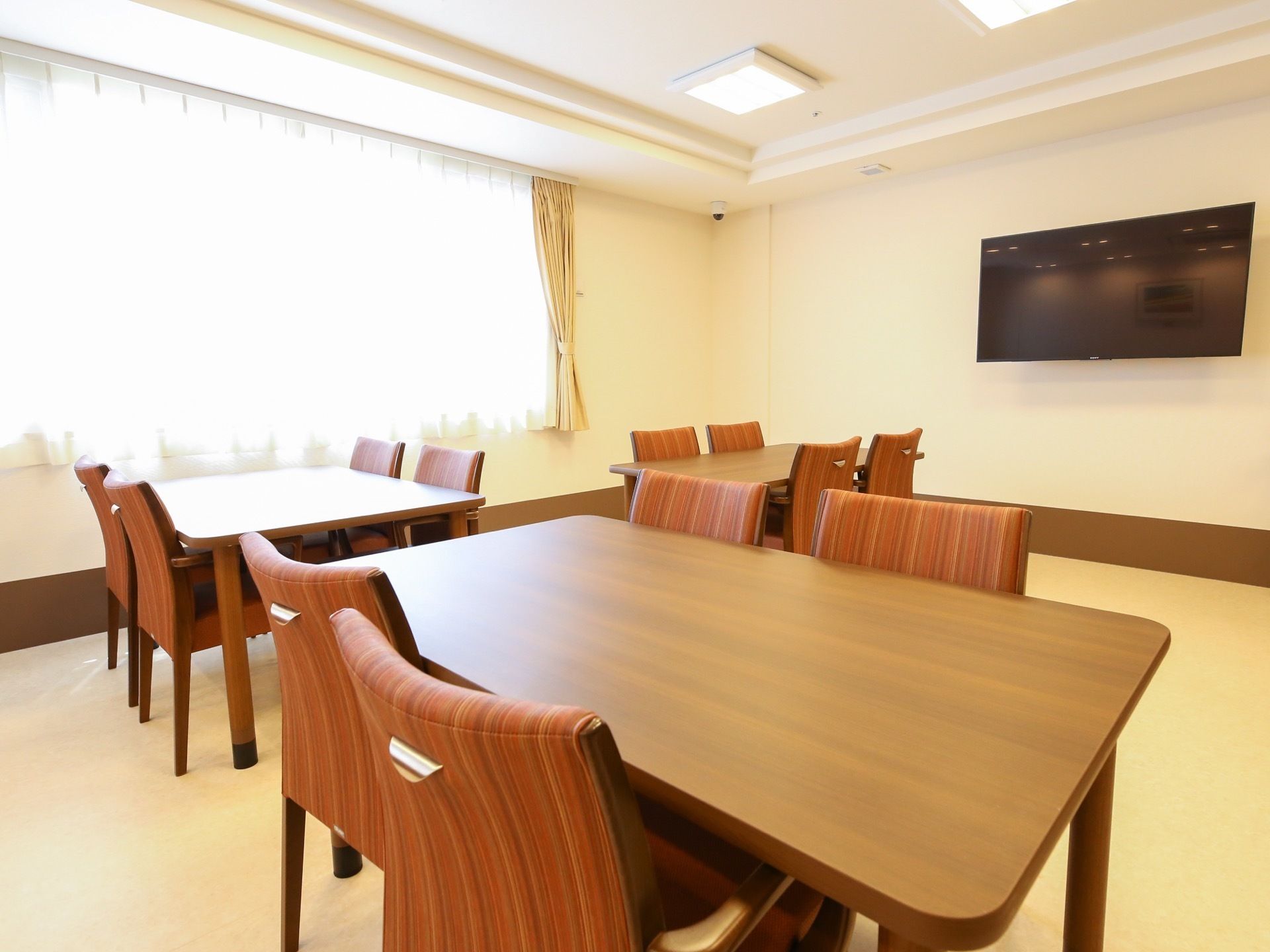 白い壁には大きな壁掛けのテレビが設置されており、複数のテーブルと椅子が置かれている空間である。窓にはカーテンがかけられている。