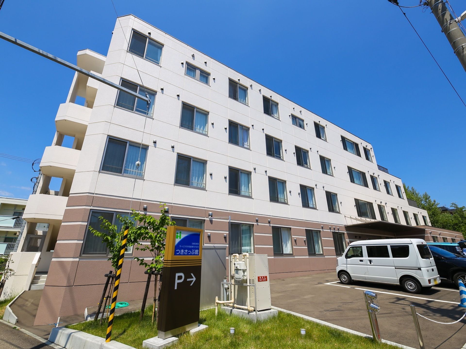 青空の下にツートンカラーの４階建ての建物が建っている。建物の端には階段が設置されており、駐車場には車が数台停まっている。