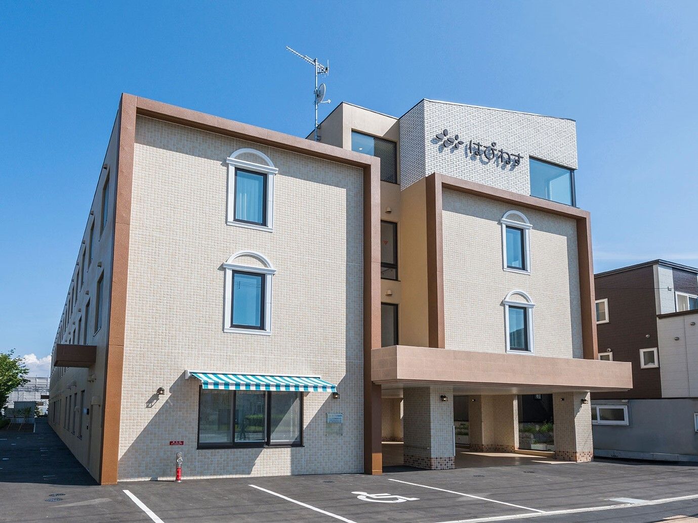 ベージュ系の3階建ての建物は日当たりのいい場所に建っていて、敷地内には駐車場があり、外壁には住宅名が書かれている。