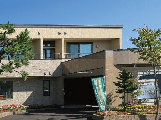 ブラウン系の外壁の2階建ての建物の周りには草木が植えられ、花壇もある。日当たりのいい場所に建っている。