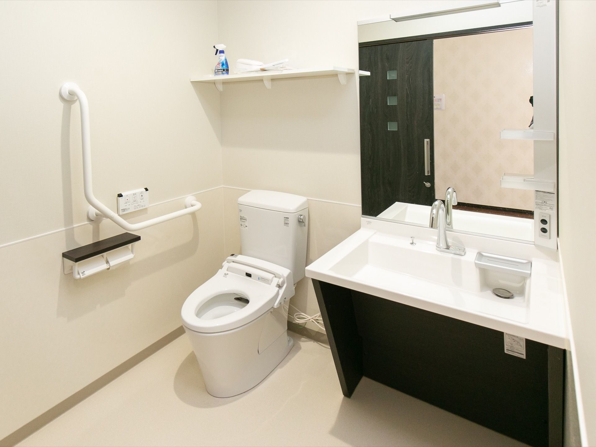 トイレは白とダークブラウンで統一されており清潔感がある。壁に手すりが備わっている。洗面台の蛇口はセンサー式である。