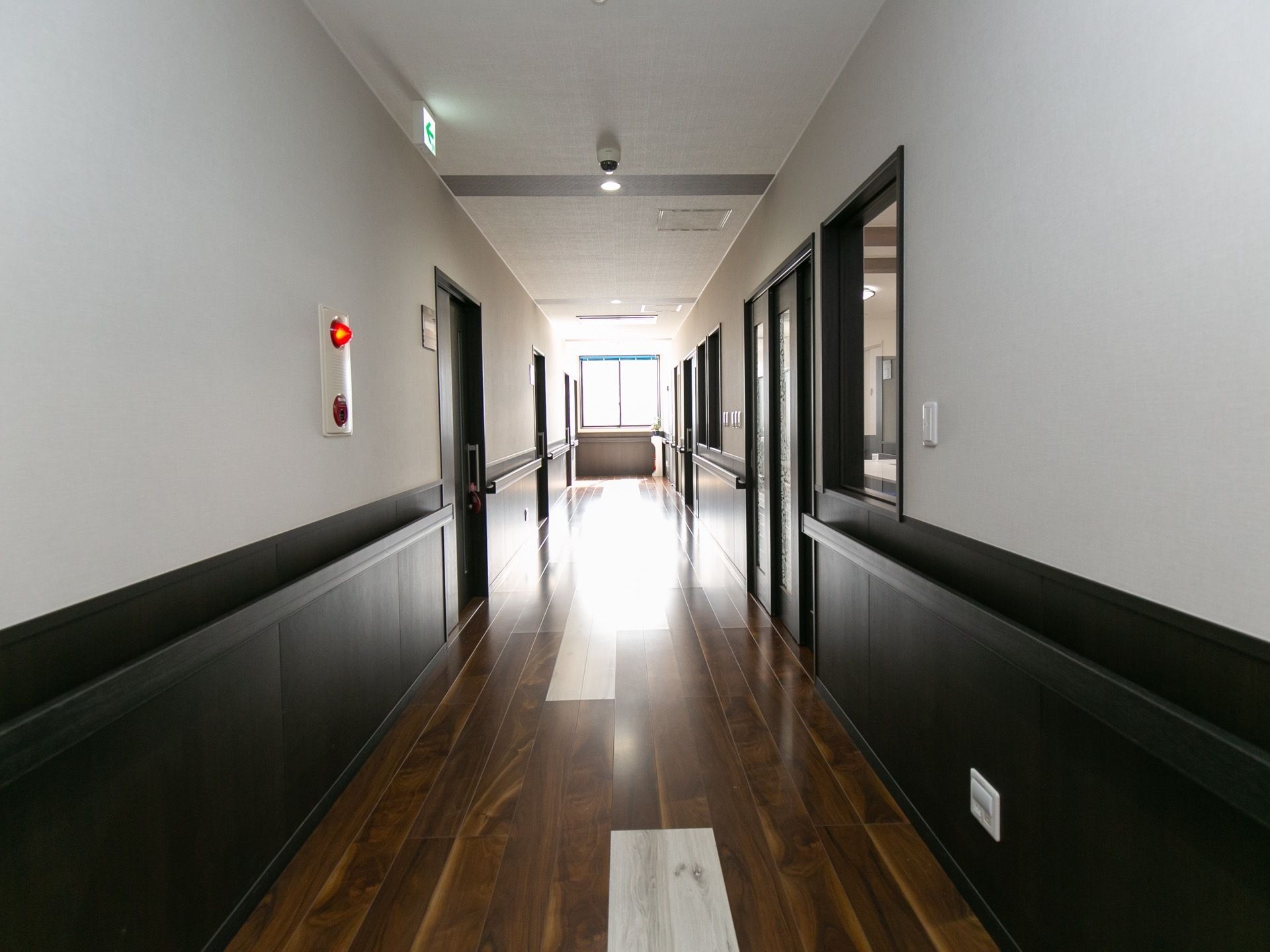 廊下には余計なものが置かれておらず、清掃が行き届いている。突き当たりに大きな窓がある。壁に火災報知器が付けられている。