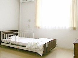 レースカーテンとカーテンが付いた大きな窓のそばには寝具付きのベッドが置かれ、緊急通報装置もある。