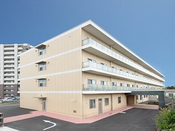 各フロアにベランダがあり、ベージュの外壁の４階建ての建物。周りには緑もある。