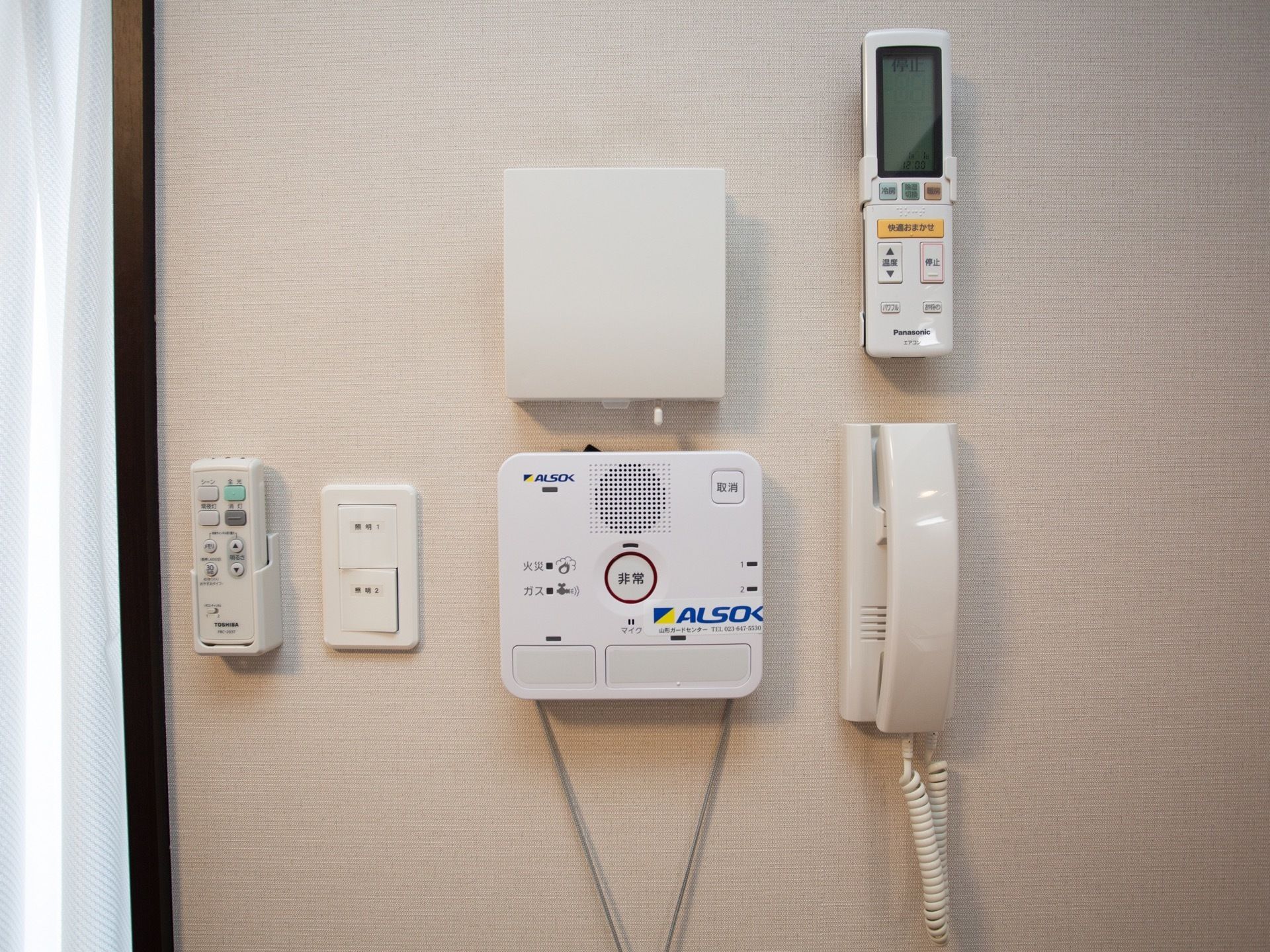 居室の壁に緊急用通報装置やエアコンのリモコン、電話機が取り付けられている。左横には、カーテンが掛けられた窓がある。