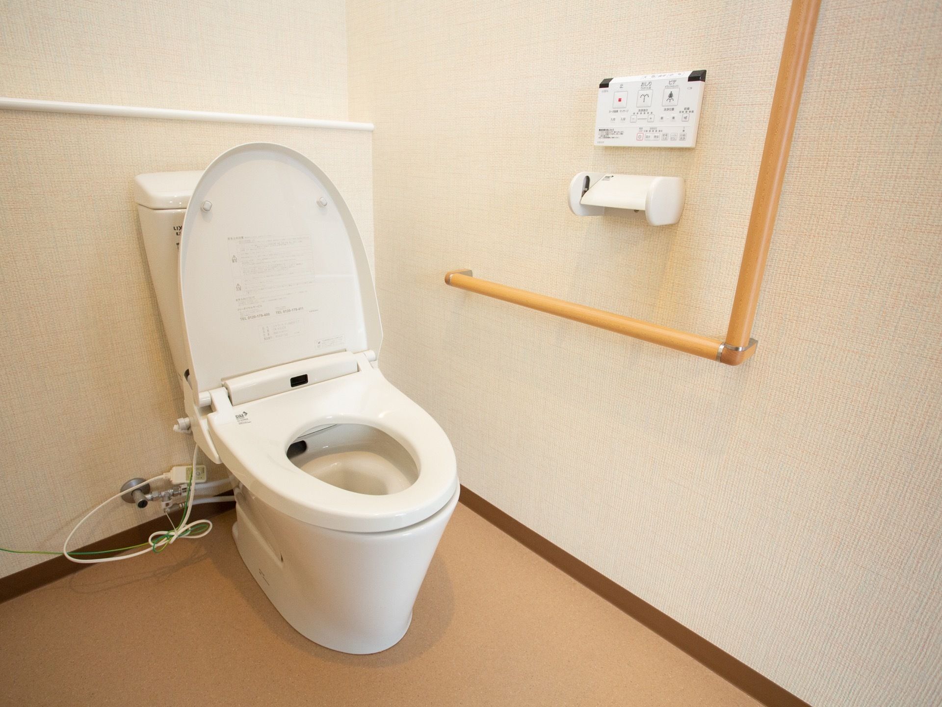 開放的な温水洗浄便座つきのトイレである。壁にはL字型の手すりとペーパーホルダーがある。