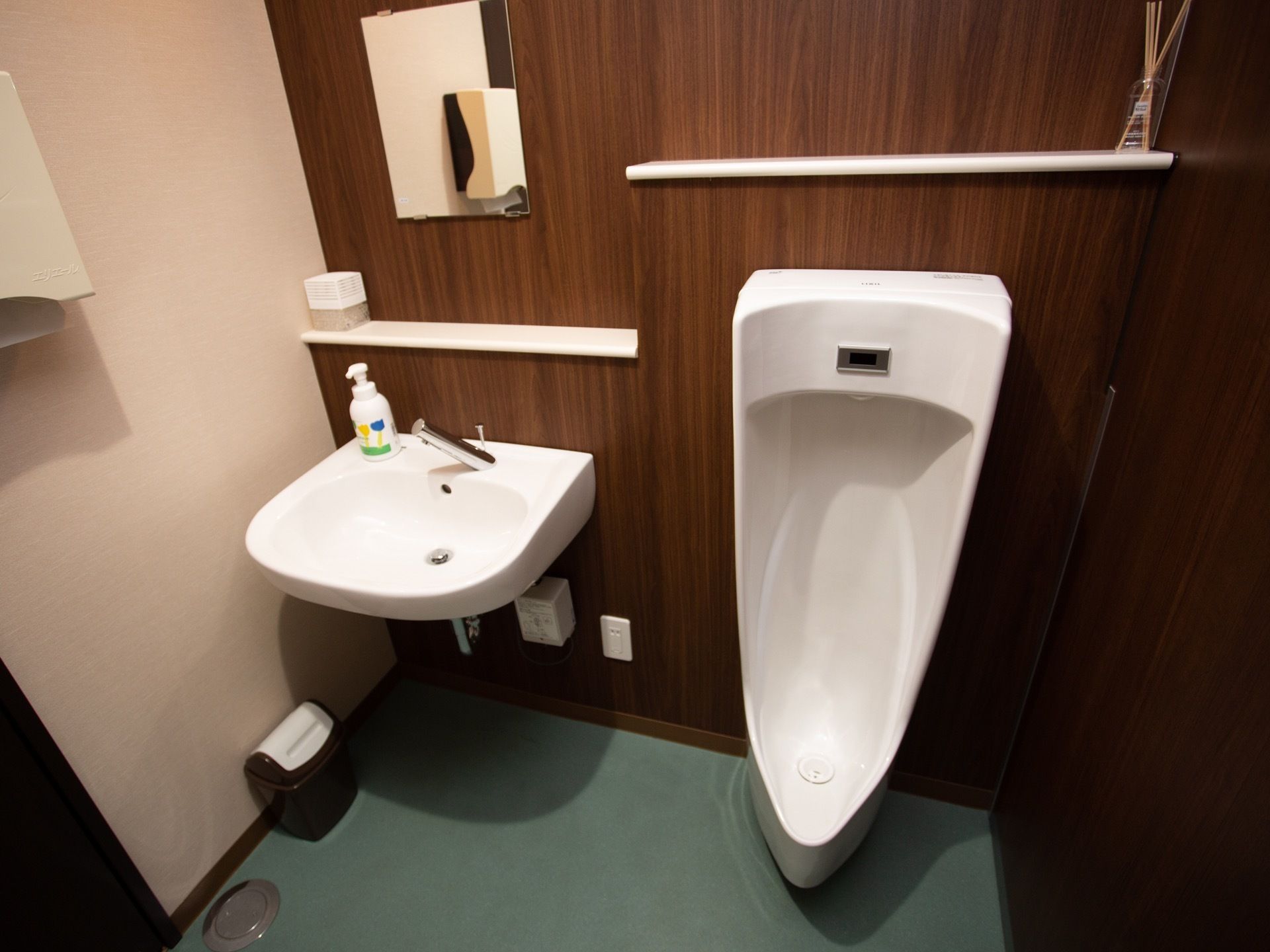 ダークトーンの落ち着いた雰囲気の男性用トイレである。男性用小便器と洗面台が隣り合わせに並んでおり、ペーパータオルも用意されている。