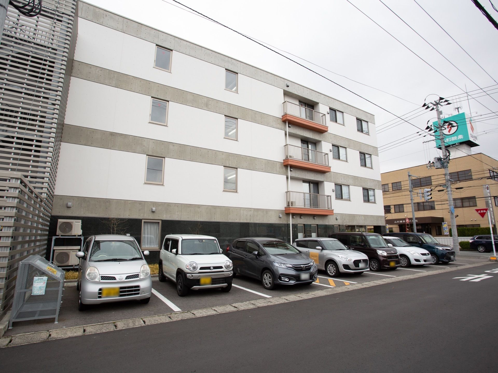 施設は1階が黒、その上階が白い壁の4階建てとなっている。敷地内には横列タイプの駐車場があり、数台の車が停められている。