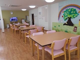 ピンクの椅子がセットされた木製のダイニングテーブルがずらりと並んだ食堂の奥にはテレビがあり、壁には木が描かれている。