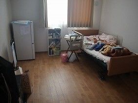 フローリングの部屋には冷蔵庫やベッドなどが設置され、カーテンが付いた大きな窓がある。