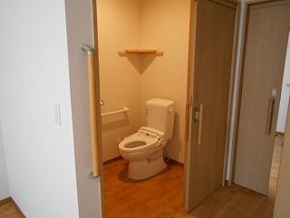 スライド式のドアが付いたトイレは広く、ふたのない便座が付いていて。壁には手すりが付いている。