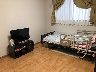 大きな窓が付いたフローリングの部屋には寝具付きのベッドやテレビが置かれ、テレビの横には時計もある。
