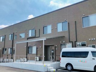 大きな屋根や手すり付きのスロープが付いた玄関がある２階建ての建物は駐車場もあり、車が止められている。