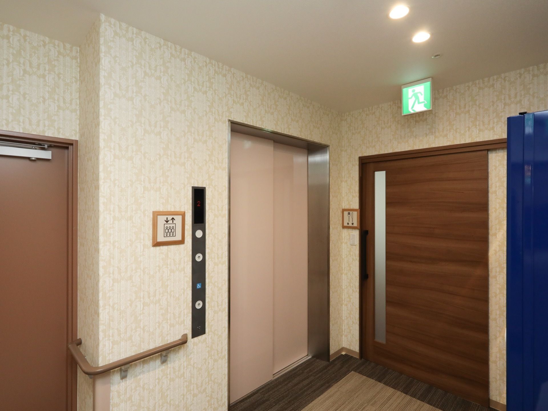 廊下にエレベーターが設置されており、扉はベージュ色になっている。天井には、緑色の非常口を表す表示が取り付けられている。
