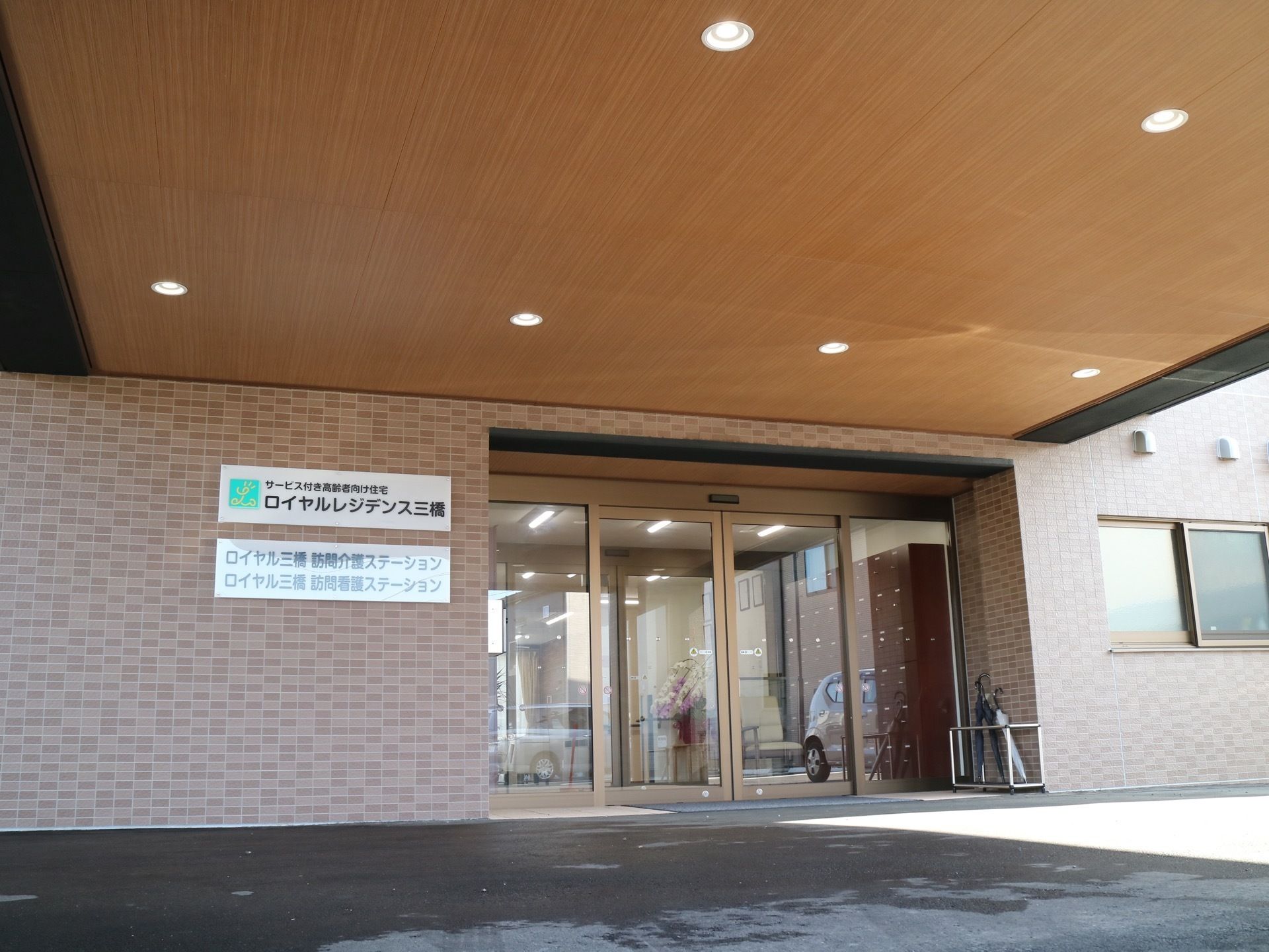 外から見た施設の正面玄関。ドアの横には看板が設置されており、ひさしとなる天井部分にはいくつか照明が取り付けられている。